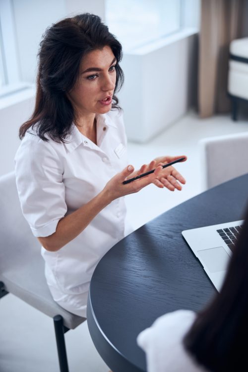 Serious middle-aged general practitioner talking to a dark-haired woman seated at the table in her office